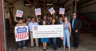 A group of SWTJC students huddle around the $380,000 contribution check from UP. Behind the check are, from left, Ken Kuwamura (in light blue), Manager-Talent Acquisition, UP; Raquel Espinoza, Senior Director-Public Affairs, UP; Esmerelda Arreola, SWTJC Diesel Technology Program Lab Assistant; Texas State Rep. Eddie Morales and his wife, Helen Morales; and Tom Blevins, Superintendent-Train Operations, UP. (Caption and Photograph Courtesy of UP)